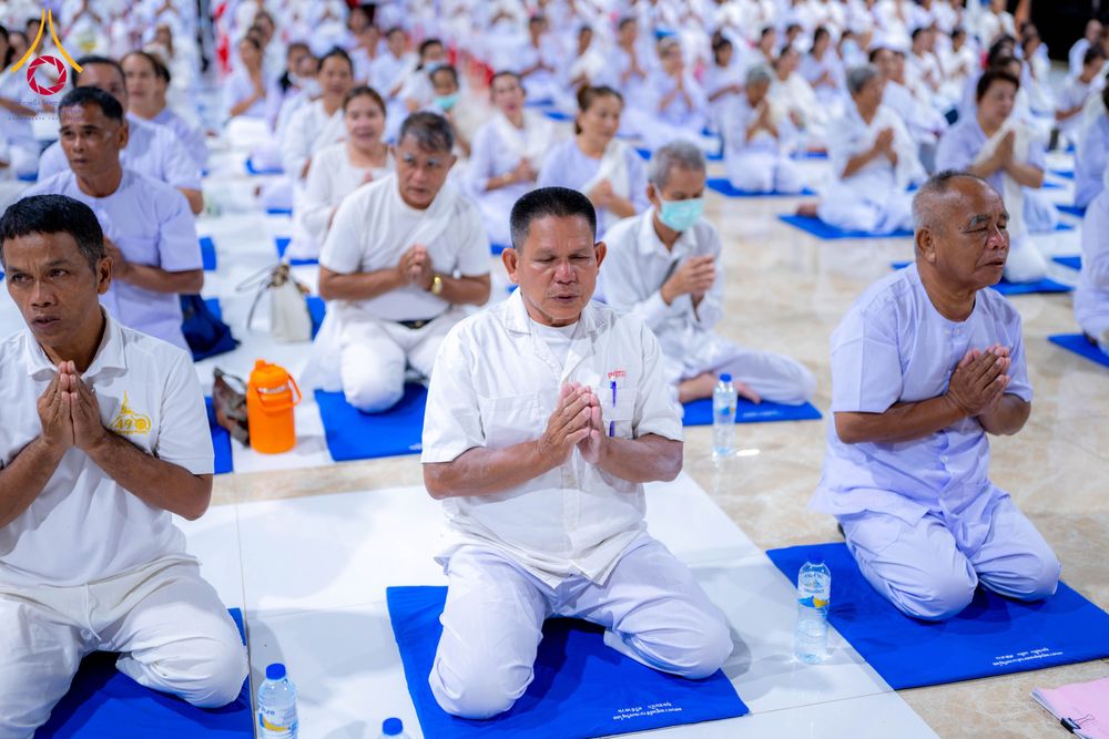 กลุ่มคนสวมชุดขาวนั่งพนมมือปฏิบัติธรรมบนเบาะรองนั่งสีน้ำเงินภายในอาคาร
