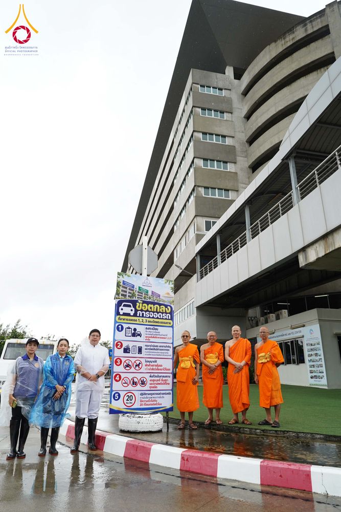 พระภิกษุสงฆ์ในจีวรสีส้มและเจ้าหน้าที่ยืนเรียงแถวหน้าป้ายกฎการจอดรถข้างอาคารสูง