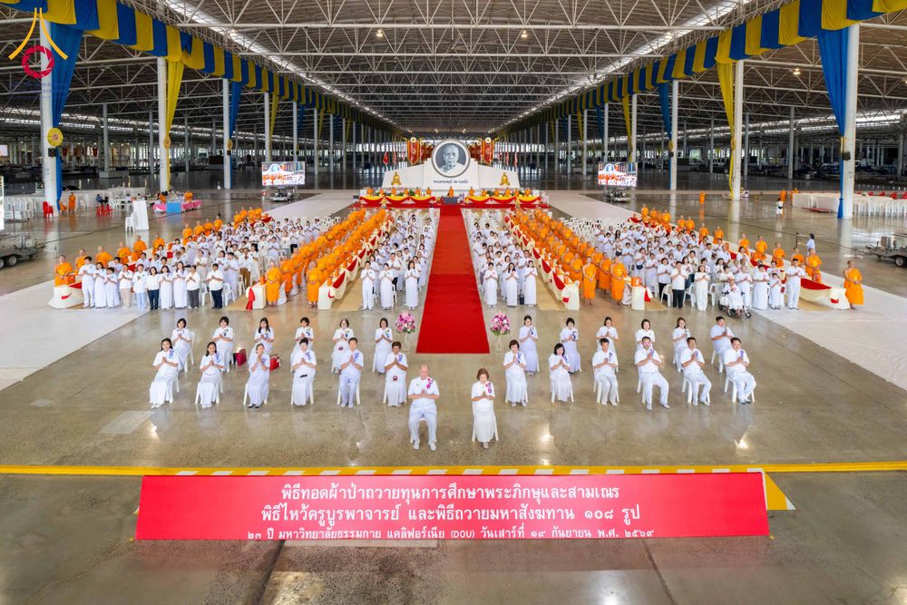 พระภิกษุและสาธุชนชุดขาวนั่งประกอบพิธีทางศาสนาภายในอาคารขนาดใหญ่ที่มีพรมแดงทอดยาวไปยังแท่นพิธี