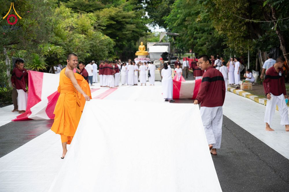 พระภิกษุและกลุ่มคนกำลังช่วยกันกางผ้าสีขาวผืนยาวบนถนนเพื่อเตรียมจัดงานพิธี