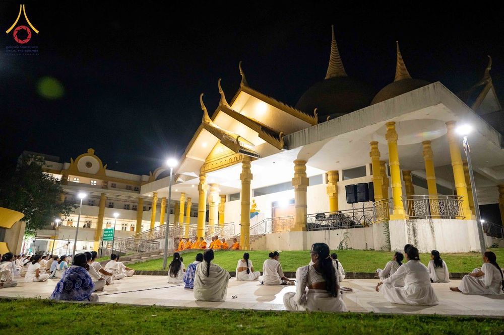 กลุ่มคนสวมชุดขาวนั่งปฏิบัติธรรมบนลานหน้าอาคารทรงไทยในยามค่ำคืน