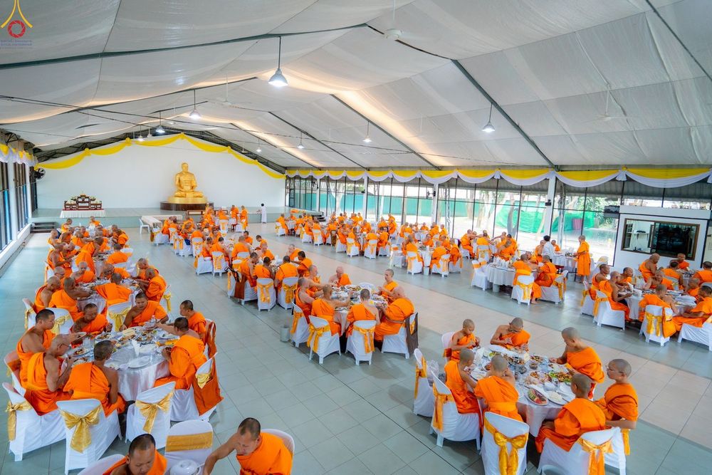 คณะสงฆ์จำนวนมากในจีวรสีส้มกำลังนั่งฉันภัตตาหารบนโต๊ะจีนภายในอาคารชั่วคราว