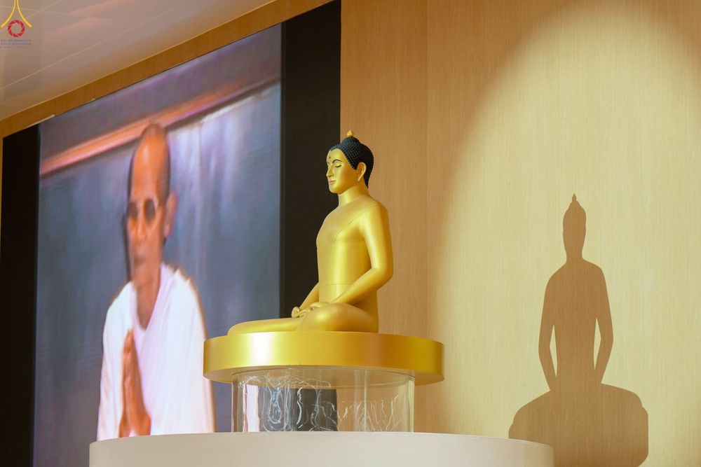 พระพุทธรูปสีทองนั่งสมาธิบนฐานใส โดยมีภาพบุคคลบนจอขนาดใหญ่ด้านหลังและเงาพระพุทธรูปบนผนัง