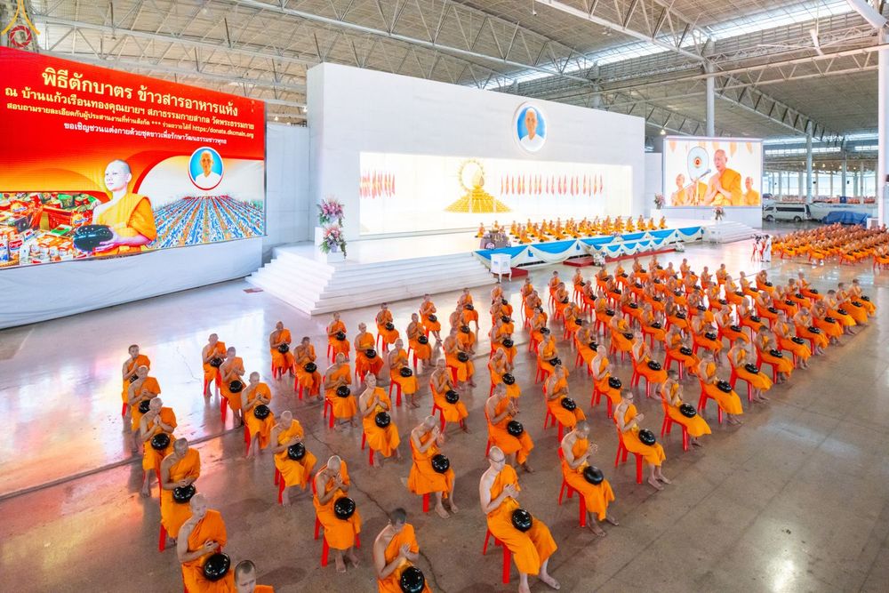 พระภิกษุสงฆ์จำนวนมากนั่งเรียงแถวบนเก้าอี้สีแดงภายในอาคารเพื่อเตรียมประกอบพิธีตักบาตร