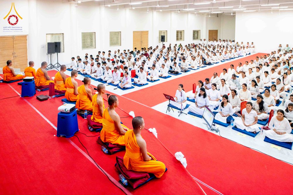 พระภิกษุสงฆ์และพุทธศาสนิกชนจำนวนมากนั่งเรียงแถวบนพรมสีแดงภายในห้องโถงเพื่อประกอบพิธีทางศาสนา