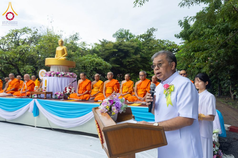 ชายสวมชุดสีขาวกำลังพูดผ่านไมโครโฟนบนเวทีที่มีพระสงฆ์นั่งอยู่ด้านหลัง