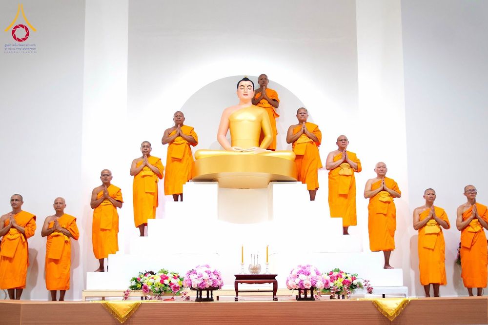 พระภิกษุสงฆ์หลายรูปยืนพนมมืออยู่บนฐานยกสูงล้อมรอบพระพุทธรูปองค์ประธานสีทองภายในอาคาร