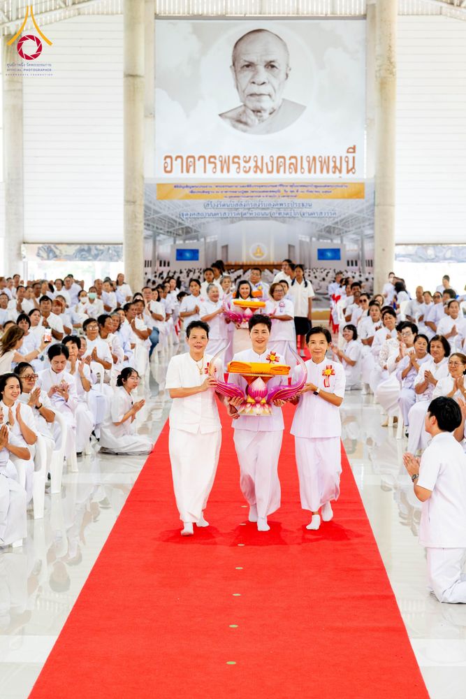 กลุ่มคนสวมชุดขาวเดินบนพรมแดงในอาคารเพื่อประกอบพิธีทางศาสนา โดยมีผู้ถือพานพุ่มดอกบัวนำหน้า