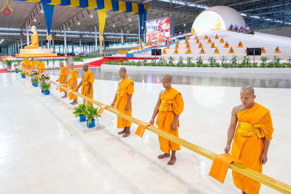 พระภิกษุและสามเณรยืนเรียงแถวถือสายผ้าป่าภายในอาคารขนาดใหญ่