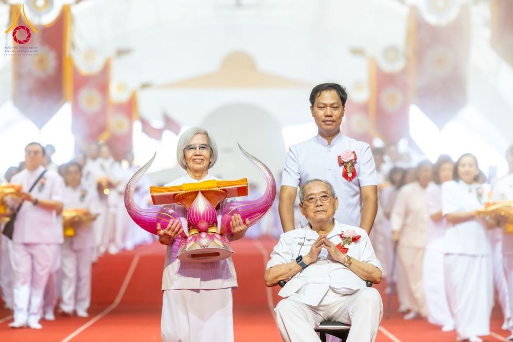 กลุ่มพุทธศาสนิกชนในชุดสีขาวร่วมเดินขบวนในพิธีทอดผ้าป่า โดยมีผู้ถือพานพุ่มผ้าไตรนำหน้า และมีผู้ชายเข็นรถเข็นที่มีชายสูงวัยนั่งพนมมืออยู่ด้านหลัง ภายในสภาธรรมกายสากล