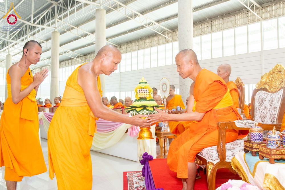 พระภิกษุสงฆ์ในจีวรสีส้มกำลังถวายพานพุ่มดอกไม้แด่พระเถระที่นั่งอยู่บนอาสนะภายในอาคาร