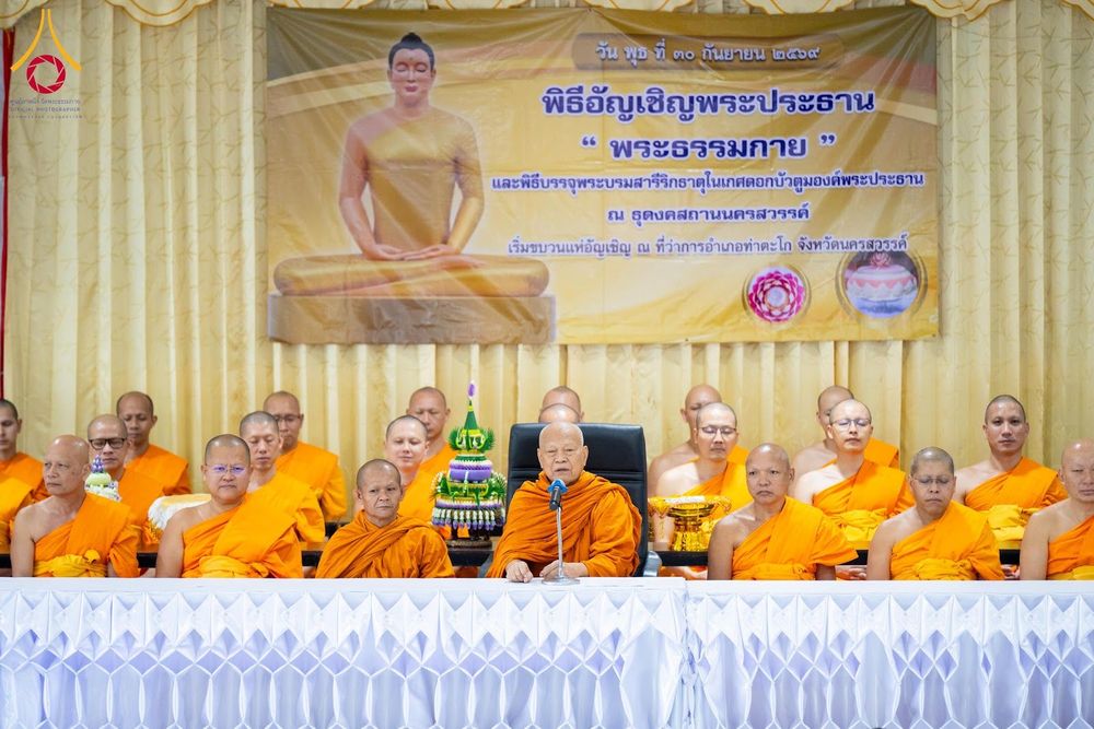 คณะสงฆ์นั่งบนเวทีในพิธีทางศาสนา ด้านหลังมีป้ายไวนิลระบุชื่อพิธีอัญเชิญพระประธานและบรรจุพระบรมสารีริกธาตุ
