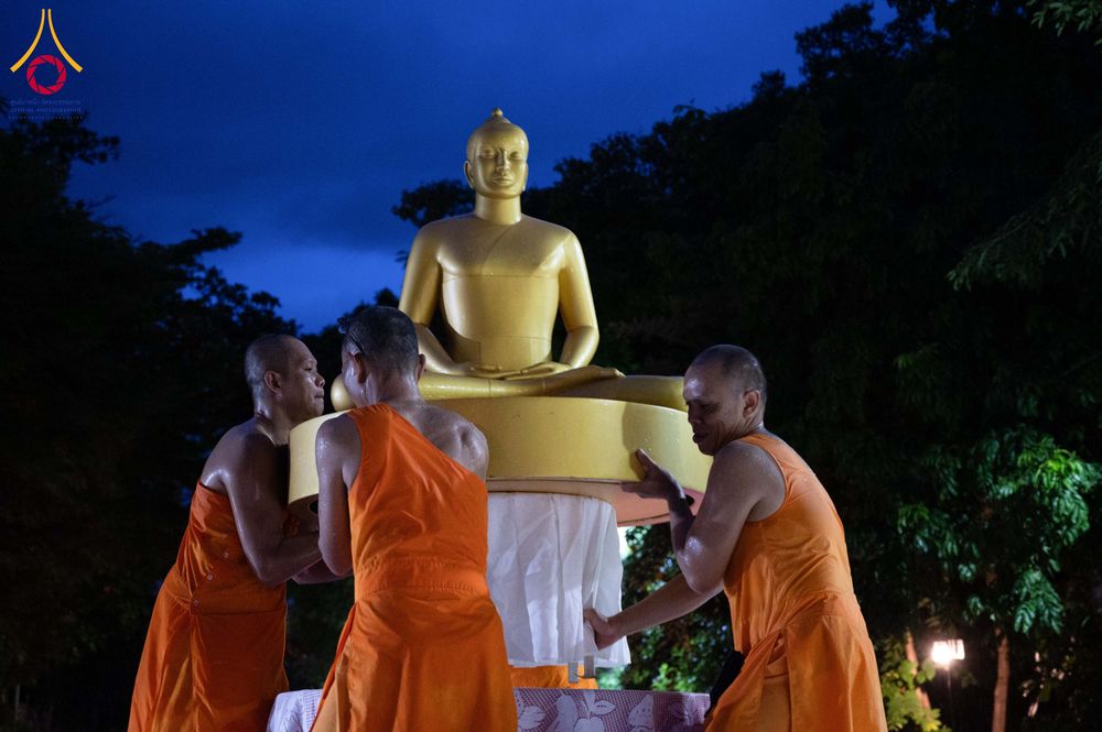 พระภิกษุ 3 รูปกำลังช่วยกันยกพระพุทธรูปสีทองในบรรยากาศยามค่ำคืน