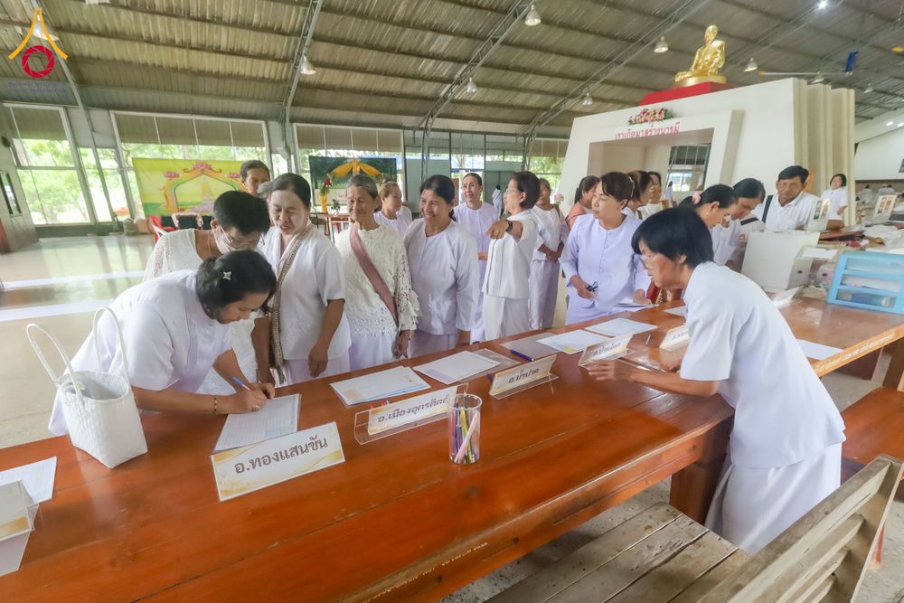 กลุ่มอุบาสิกาในชุดสีขาวกำลังเขียนเอกสารลงทะเบียนบนโต๊ะไม้ภายในอาคาร