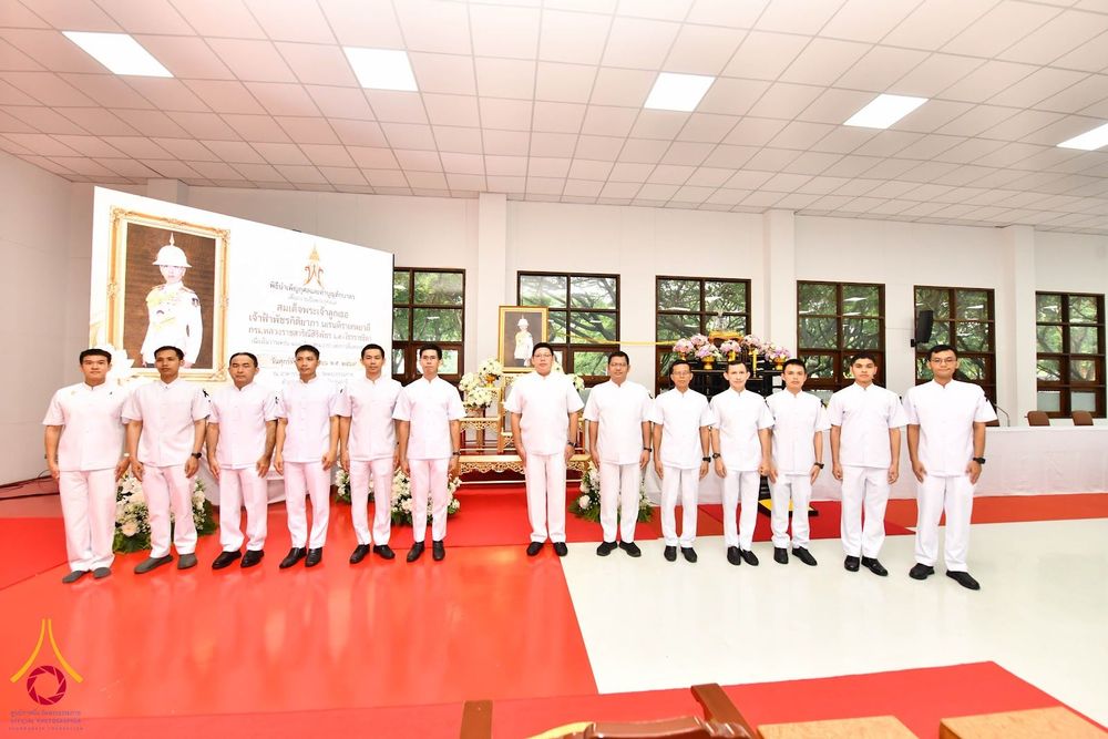 กลุ่มชายในชุดสีขาวสุภาพยืนเรียงแถวหน้ากระดานในห้องโถงที่มีการจัดโต๊ะหมู่บูชาและป้ายพิธีบำเพ็ญกุศล