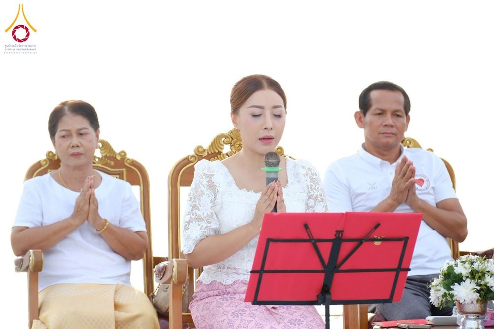 หญิงสาวถือไมโครโฟนและพนมมือประกอบพิธีร่วมกับผู้ร่วมงานชายและหญิง