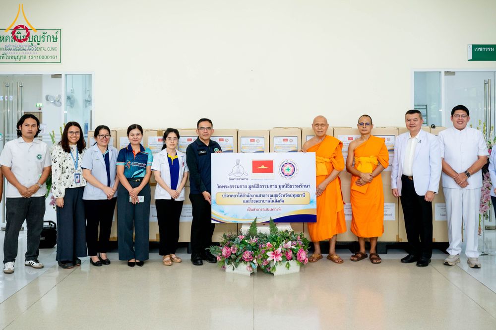 กลุ่มบุคคลและพระภิกษุสงฆ์ยืนเรียงแถวหน้ากล่องพัสดุจำนวนมาก พร้อมป้ายข้อความระบุถึงการบริจาคยาและเวชภัณฑ์