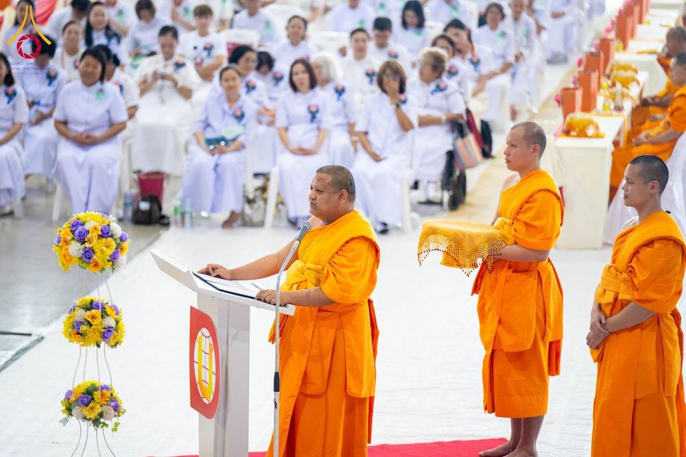 พระภิกษุสงฆ์กำลังกล่าวธรรมบรรยายที่โพเดียมในพิธีทางศาสนาที่มีสาธุชนชุดขาวนั่งร่วมงาน