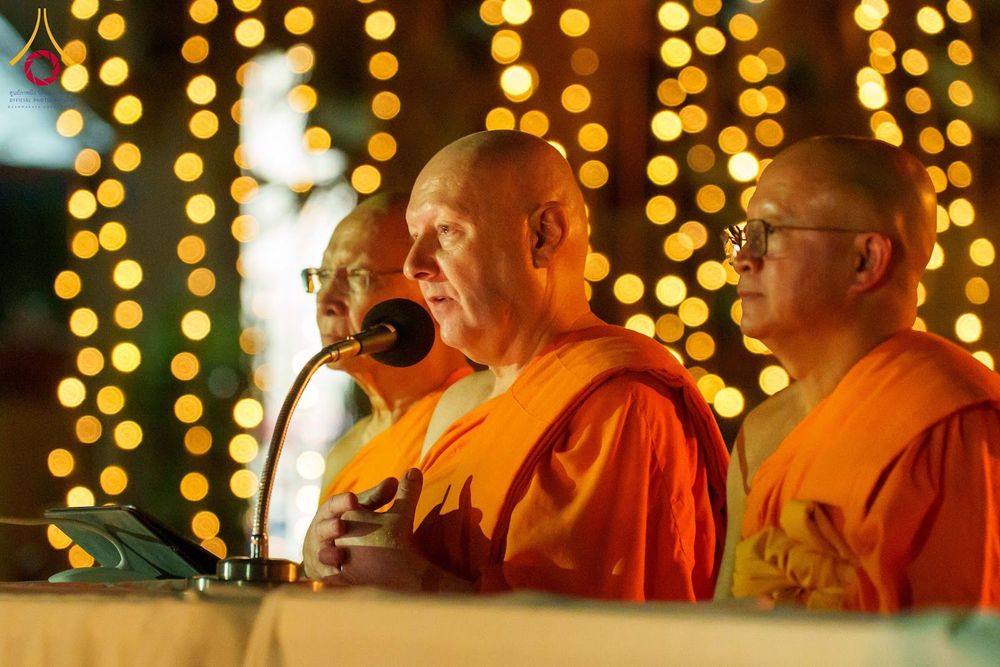 พระภิกษุสงฆ์สามรูปกำลังสนทนาธรรมหน้าไมโครโฟนท่ามกลางแสงไฟประดับในยามค่ำคืน