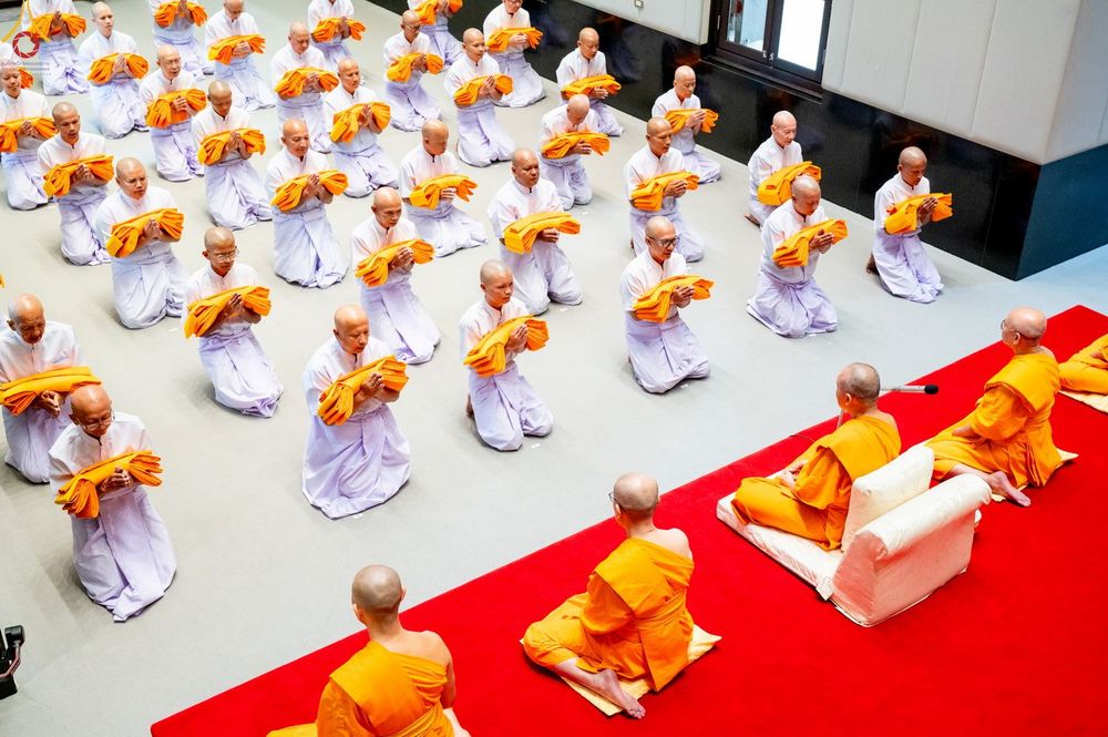 กลุ่มผู้เข้าร่วมพิธีบรรพชาอุปสมบทในชุดสีขาวนั่งพนมมือถือผ้าไตรจีวร โดยมีพระภิกษุนั่งอยู่บนพรมแดงด้านหน้า