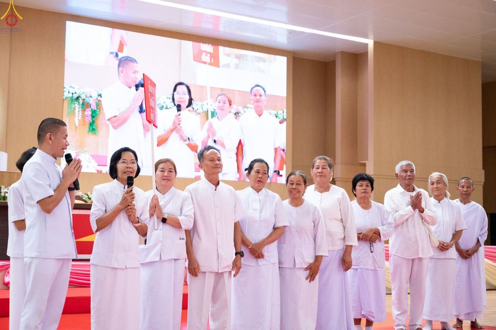 กลุ่มคนในชุดสีขาวกำลังยืนบนเวทีในงานสัมมนา โดยมีผู้ชายถือไมโครโฟนพูดและมีจอภาพขนาดใหญ่ด้านหลัง