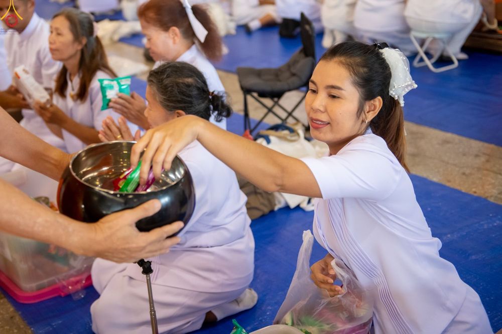 หญิงสาวสวมชุดขาวกำลังหย่อนสิ่งของลงในบาตรพระในพิธีตักบาตรท่ามกลางพุทธศาสนิกชนที่นั่งเรียงแถว