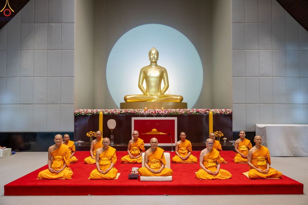 พระภิกษุสงฆ์นั่งสมาธิบนอาสนะสีแดงเบื้องหน้าพระพุทธรูปสีทองภายในอาคาร