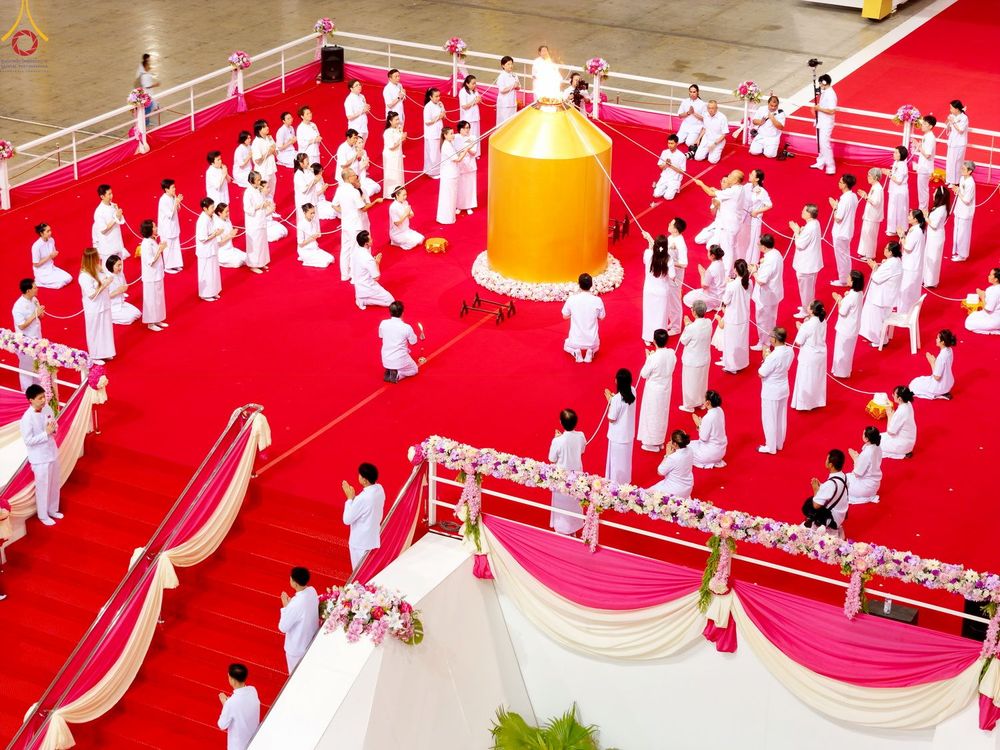 ผู้คนสวมชุดสีขาวนั่งพนมมือล้อมรอบแท่นสีทองที่มีเปลวไฟในพิธีทางศาสนา