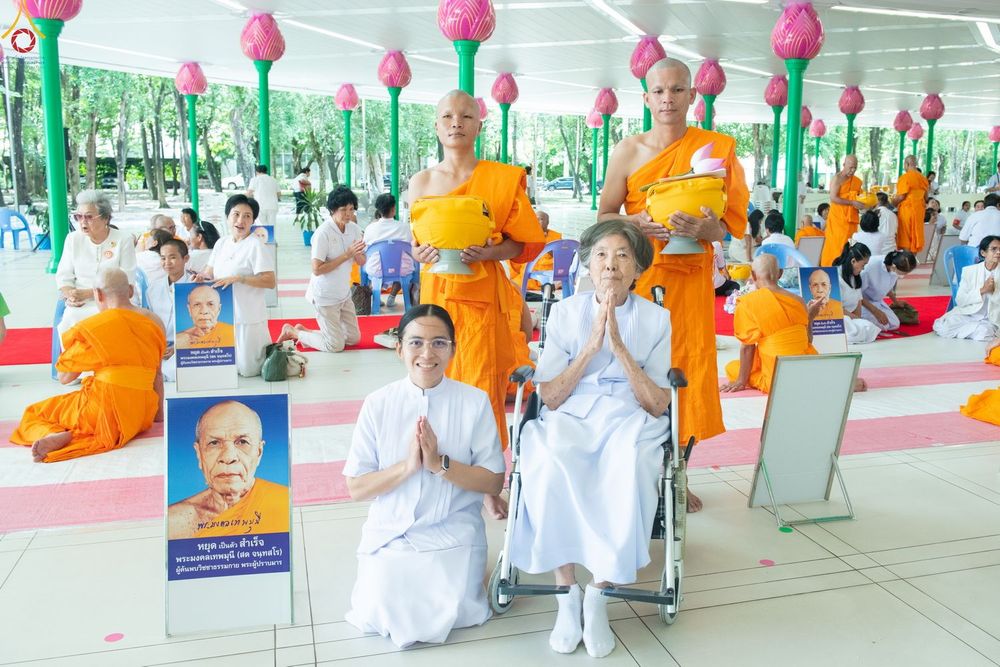 พระภิกษุสงฆ์และอุบาสิกาในชุดขาวร่วมพิธีบรรพชาอุปสมบทหมู่ภายในอาคารที่มีการตกแต่งด้วยโคมไฟรูปดอกบัว