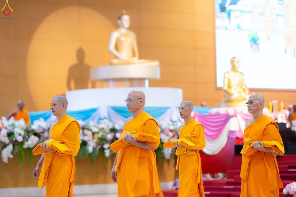พระภิกษุสงฆ์หลายรูปถือผ้าไตรจีวรเดินในพิธีทางศาสนาภายในอาคารที่มีพระพุทธรูปเป็นฉากหลัง