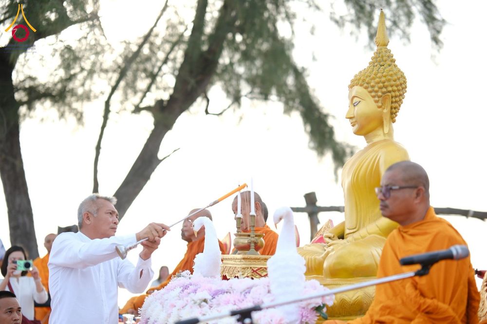 ชายสวมเสื้อสีขาวกำลังจุดเทียนบูชาหน้าพระพุทธรูปสีทองที่มีพระสงฆ์นั่งอยู่ใกล้ๆ