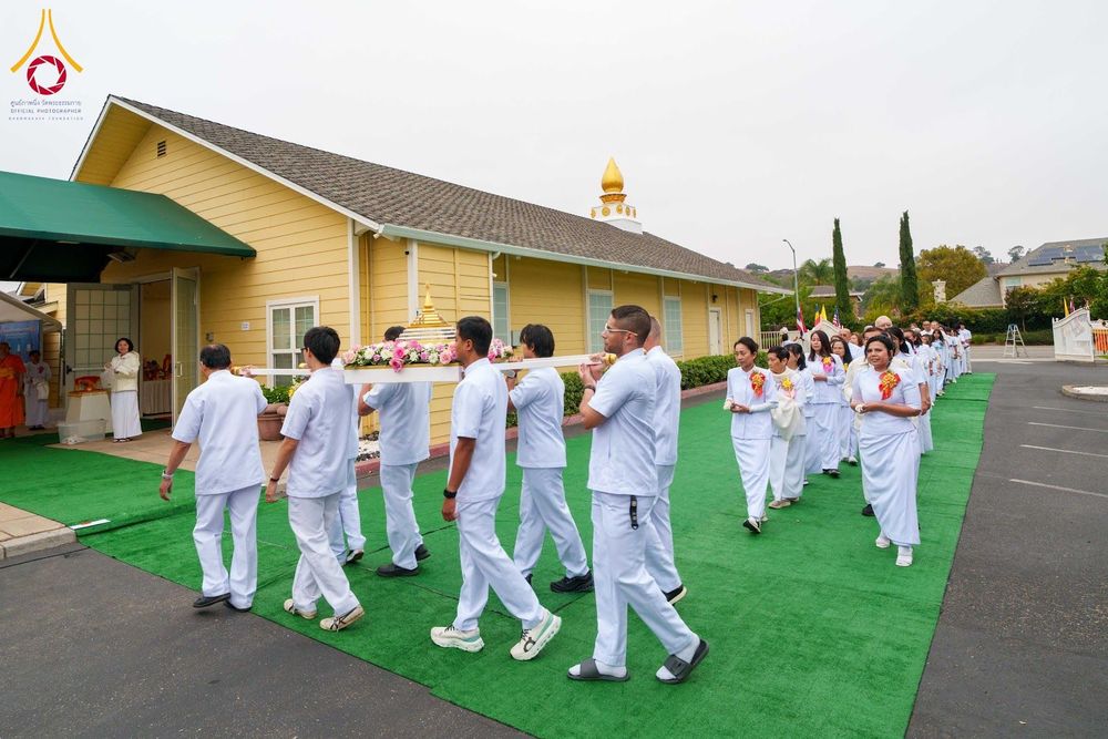 กลุ่มคนสวมชุดสีขาวเดินเป็นแถวอัญเชิญบุษบกที่มีพระบรมสารีริกธาตุบนพรมสีเขียวหน้าอาคาร