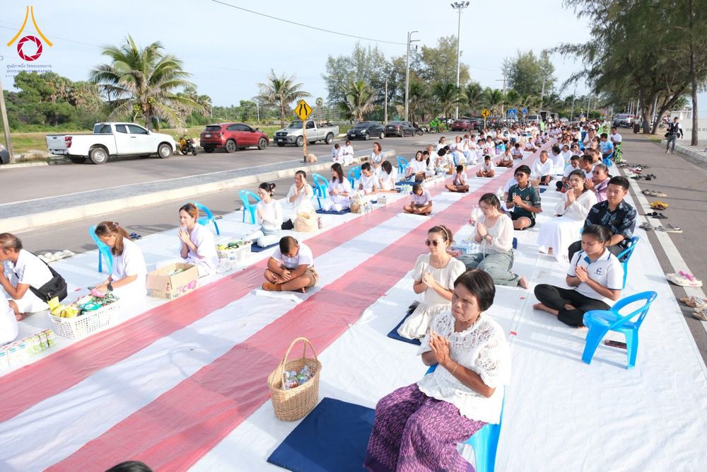 กลุ่มประชาชนนั่งพนมมือบนเสื่อสีขาวแดงริมถนนเลียบชายหาดเพื่อรอตักบาตร
