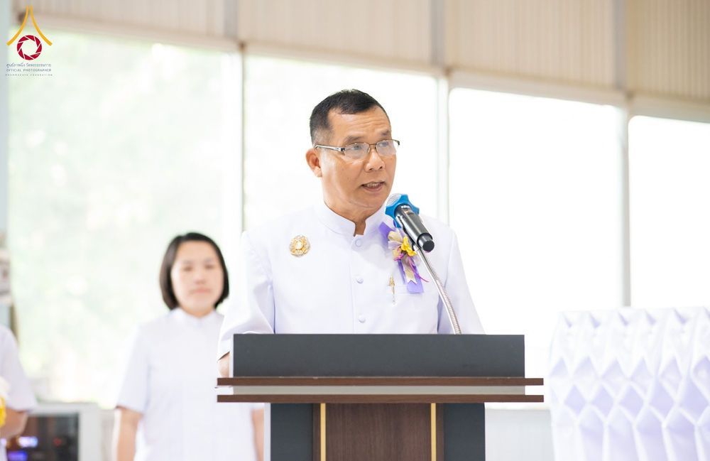 ชายสวมชุดขาวกำลังพูดผ่านไมโครโฟนที่โพเดียมในงานพิธี