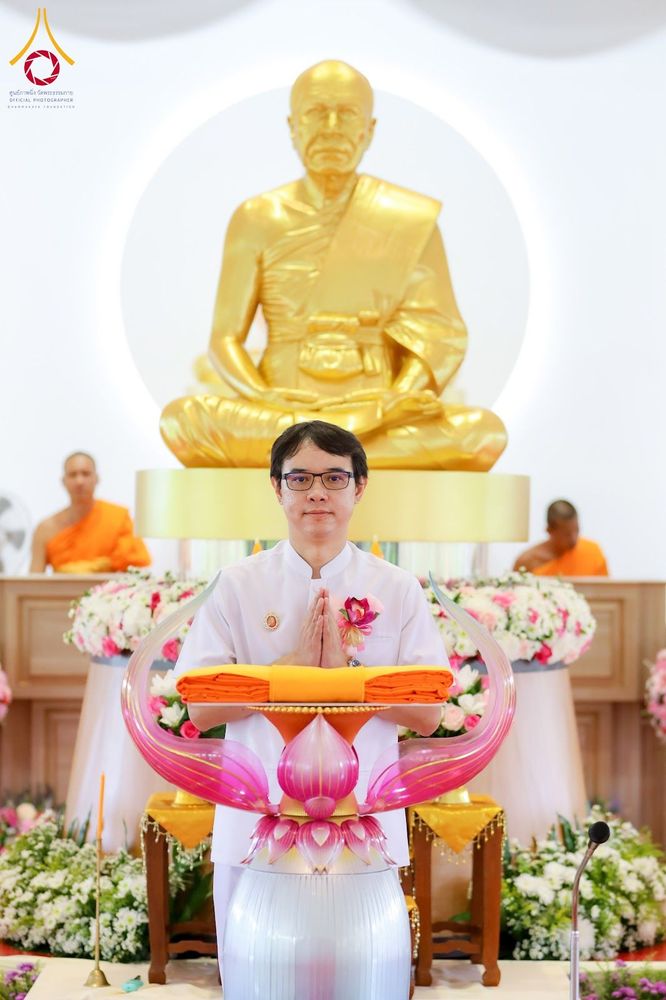 ชายสวมชุดขาวประนมมือถือผ้าไตรสีเหลืองทองหน้าพระพุทธรูปสีทองและพระสงฆ์
