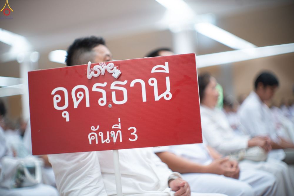 ป้ายสีแดงเขียนว่า อุดรธานี คันที่ 3 ในมือของผู้เข้าร่วมงานที่สวมชุดสีขาว