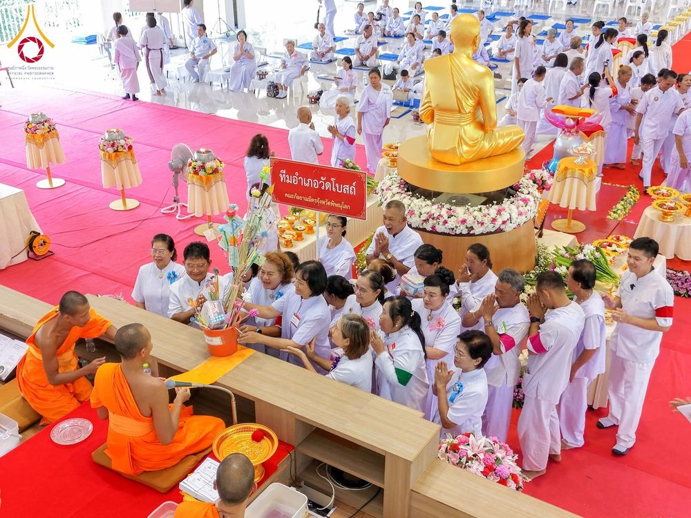 กลุ่มพุทธศาสนิกชนในชุดสีขาวร่วมกิจกรรมทางศาสนาภายในอาคาร โดยมีพระภิกษุสงฆ์และรูปปั้นพระพุทธรูปสีทองเป็นจุดศูนย์กลาง