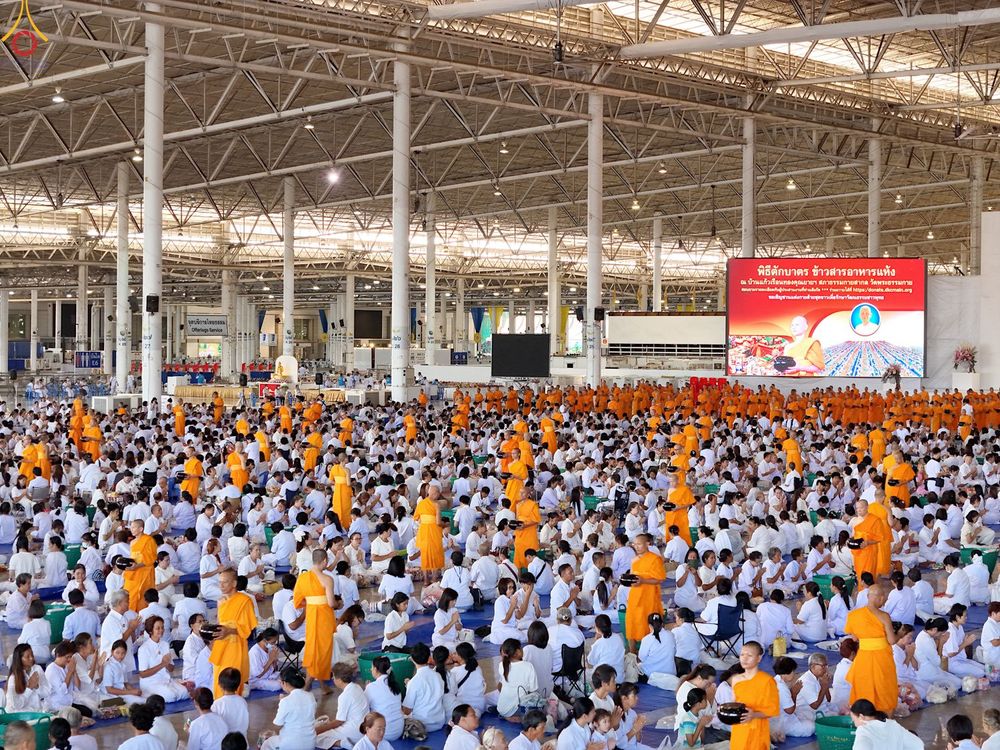 พุทธศาสนิกชนสวมชุดขาวนั่งร่วมพิธีตักบาตรข้าวสารอาหารแห้งภายในอาคารขนาดใหญ่ที่มีพระภิกษุสงฆ์เดินรับบิณฑบาต