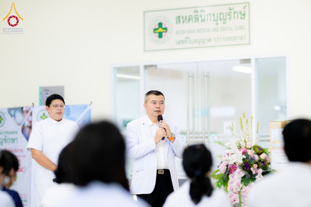 ชายสวมเสื้อกาวน์สีขาวกำลังพูดผ่านไมโครโฟนในพิธีมอบยา ณ สหคลินิกบุญรักษา