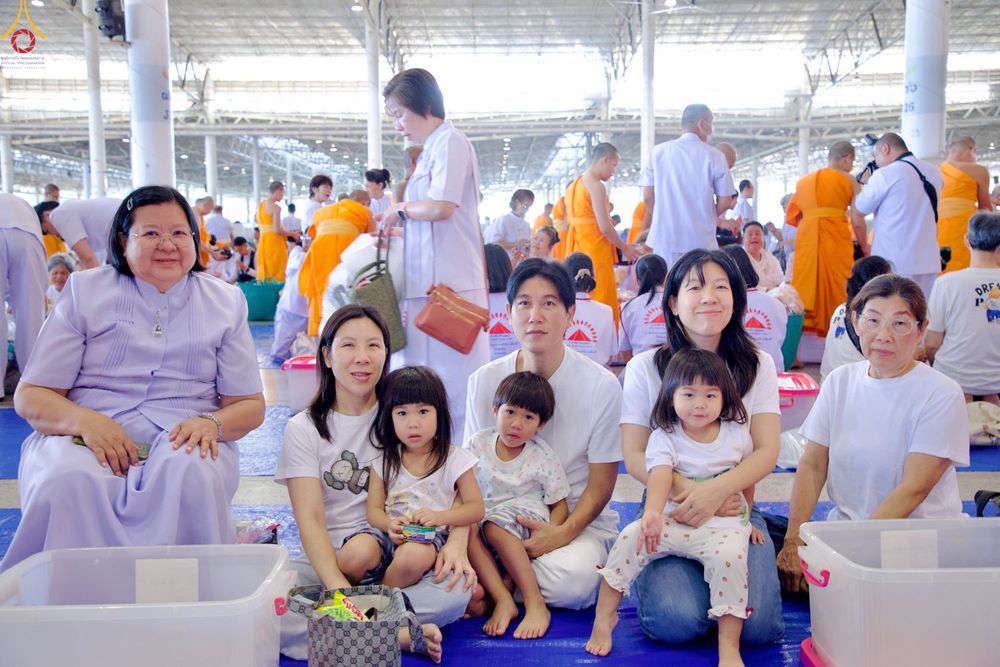 ครอบครัวชาวไทยหลายคนนั่งรวมกลุ่มกันในงานพิธีตักบาตรท่ามกลางผู้คนและพระภิกษุสงฆ์