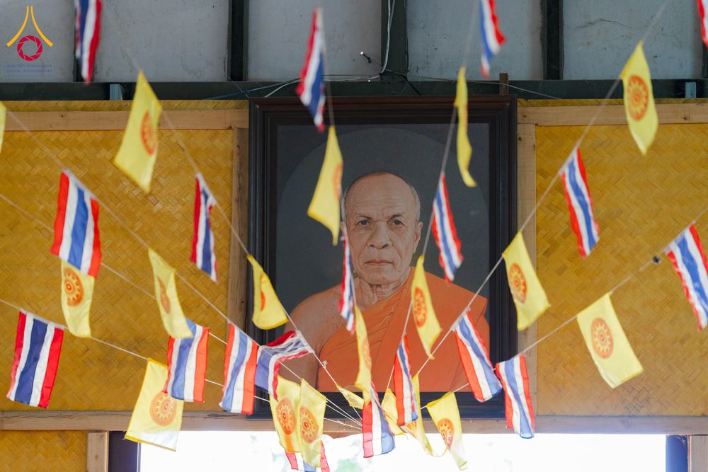 ภาพวาดพระภิกษุสงฆ์ในกรอบไม้ มีธงชาติไทยและธงธรรมจักรห้อยประดับอยู่ด้านหน้าบนพื้นหลังผนังไม้ไผ่