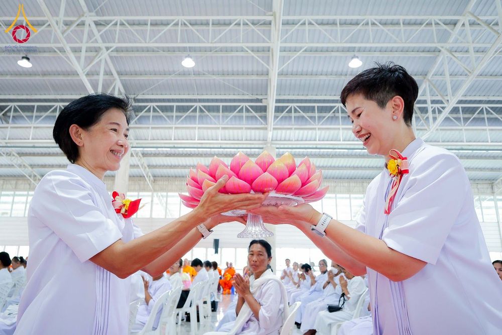 หญิงสองคนสวมชุดขาวกำลังประคองพานดอกบัวสีชมพูในพิธีทางศาสนา