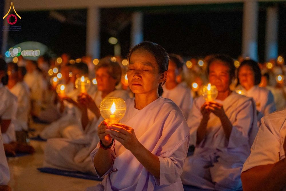 อุบาสิกาแก้วสวมชุดสีขาวนั่งหลับตาถือประทีปเทียนในมือท่ามกลางกลุ่มผู้เข้าร่วมพิธี