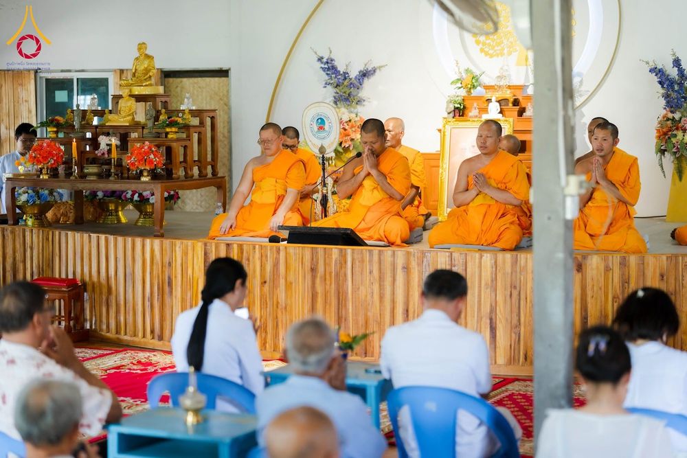 พระภิกษุสงฆ์ในจีวรสีส้มกำลังประกอบพิธีทางศาสนาบนเวทีไม้ โดยมีสาธุชนนั่งพนมมือร่วมพิธีอยู่เบื้องหน้า