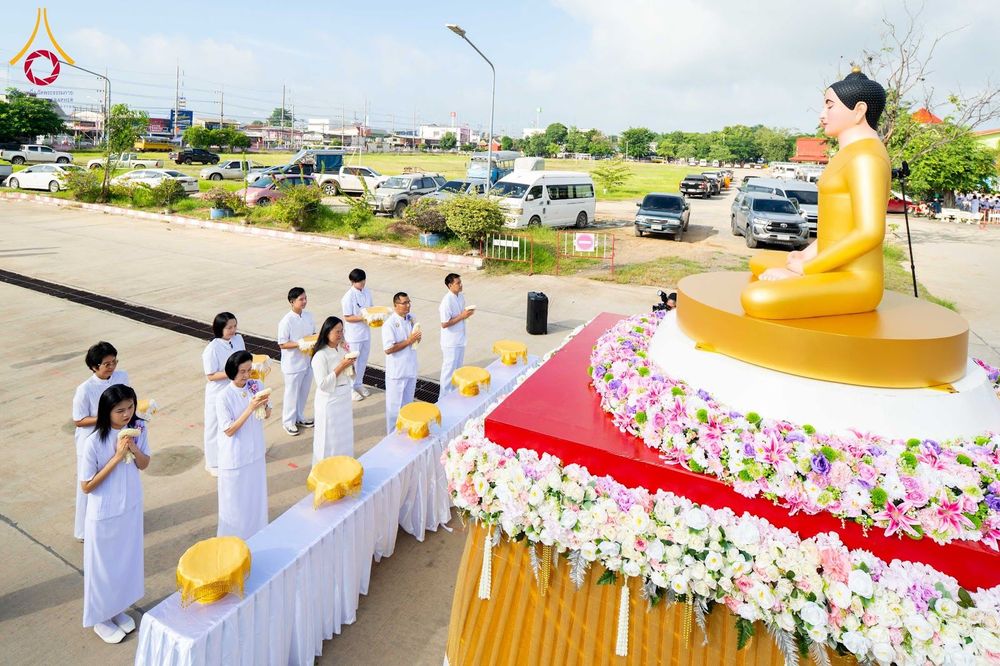 กลุ่มคนในชุดขาวกำลังประกอบพิธีทางศาสนาหน้าพระพุทธรูปสีทองที่ประดับด้วยดอกไม้กลางแจ้ง