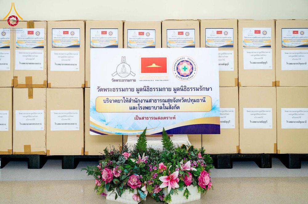 กล่องบรรจุยาจำนวนมากวางเรียงซ้อนกันพร้อมป้ายโครงการบริจาคยาเพื่อสาธารณสงเคราะห์โดยวัดพระธรรมกายและมูลนิธิที่เกี่ยวข้อง