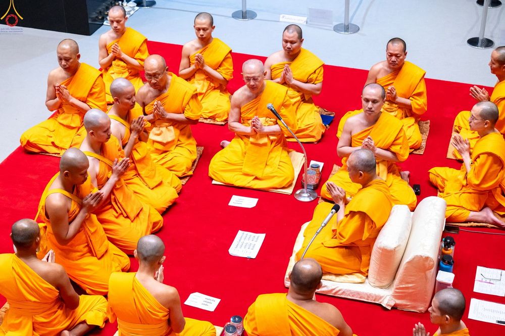 พระภิกษุสงฆ์จำนวนมากในจีวรสีเหลืองทองกำลังนั่งพนมมือประกอบพิธีกรรมทางศาสนาบนพรมสีแดง