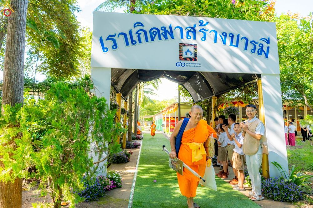 พระภิกษุสงฆ์เดินผ่านซุ้มประตูทางเข้างานบุญที่มีข้อความภาษาไทยและพุทธศาสนิกชนยืนรอต้อนรับ