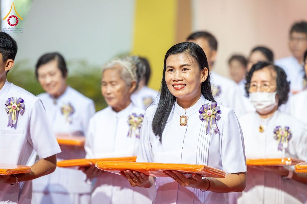 หญิงไทยสวมชุดสีขาวถือผ้าไตรจีวรสีส้มในพิธีทางศาสนาท่ามกลางพุทธศาสนิกชนท่านอื่น
