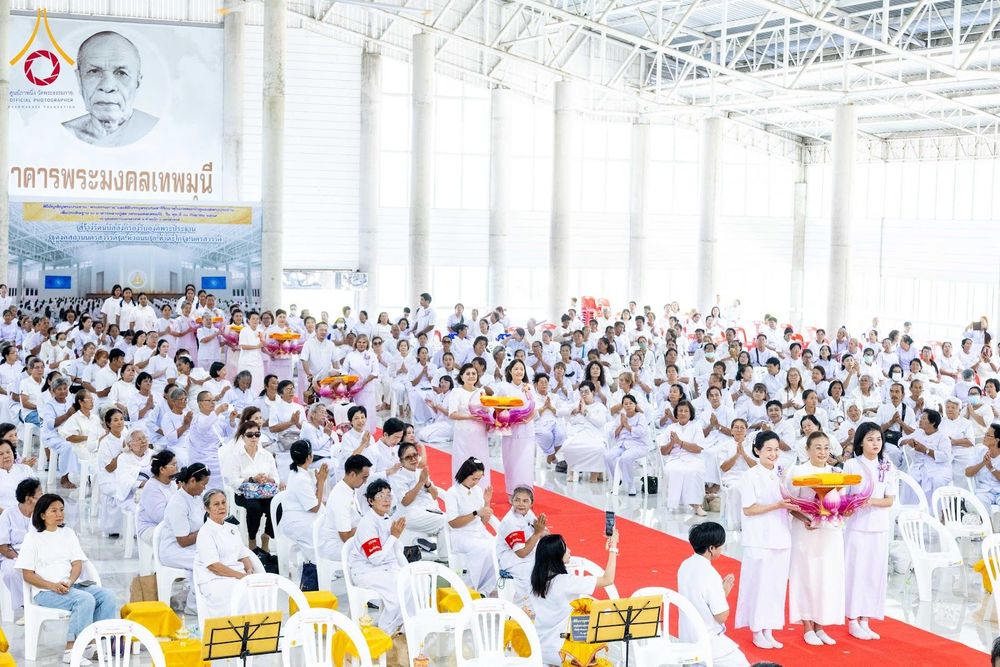 กลุ่มพุทธศาสนิกชนสวมชุดขาวนั่งและยืนร่วมพิธีทางศาสนาภายในอาคารโถงขนาดใหญ่